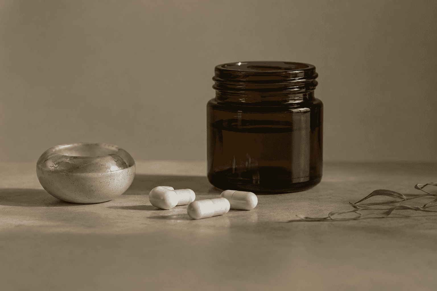 White capsules beside a small marble bowl and an amber glass jar, with olive leaves at the edge.